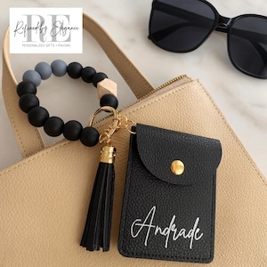 May include: A black wristlet keychain with a black tassel and gold hardware, paired with a black cardholder with the name "Andrade" in white script. The keychain features black, gray, and wooden beads.