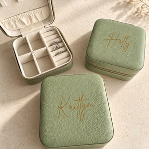 May include: Three sage green square jewelry boxes with a textured finish. One box is open, revealing a cream-colored interior with compartments and three rings. Two closed boxes feature gold script text reading Holly and Kaitlyn.