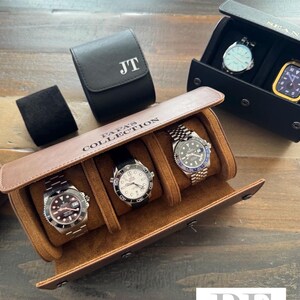 May include: A brown leather watch roll with three slots for watches. The watch roll has the text "Papa's Collection" embossed on the front. There are three watches in the slots, one with a black face, one with a white face, and one with a blue face. The watch roll is on a wooden surface.