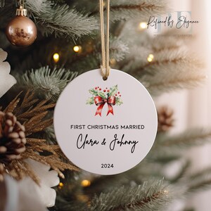 May include: White round Christmas ornament with a red bow and green holly leaves. The ornament says "First Christmas Married Clara & Johnny 2024".