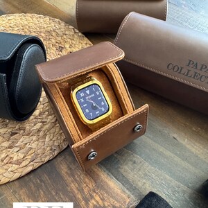 May include: A brown leather watch box with a gold watch inside. The box has a flap that closes with a snap. The watch has a black face and gold hands. The box is sitting on a wooden surface.