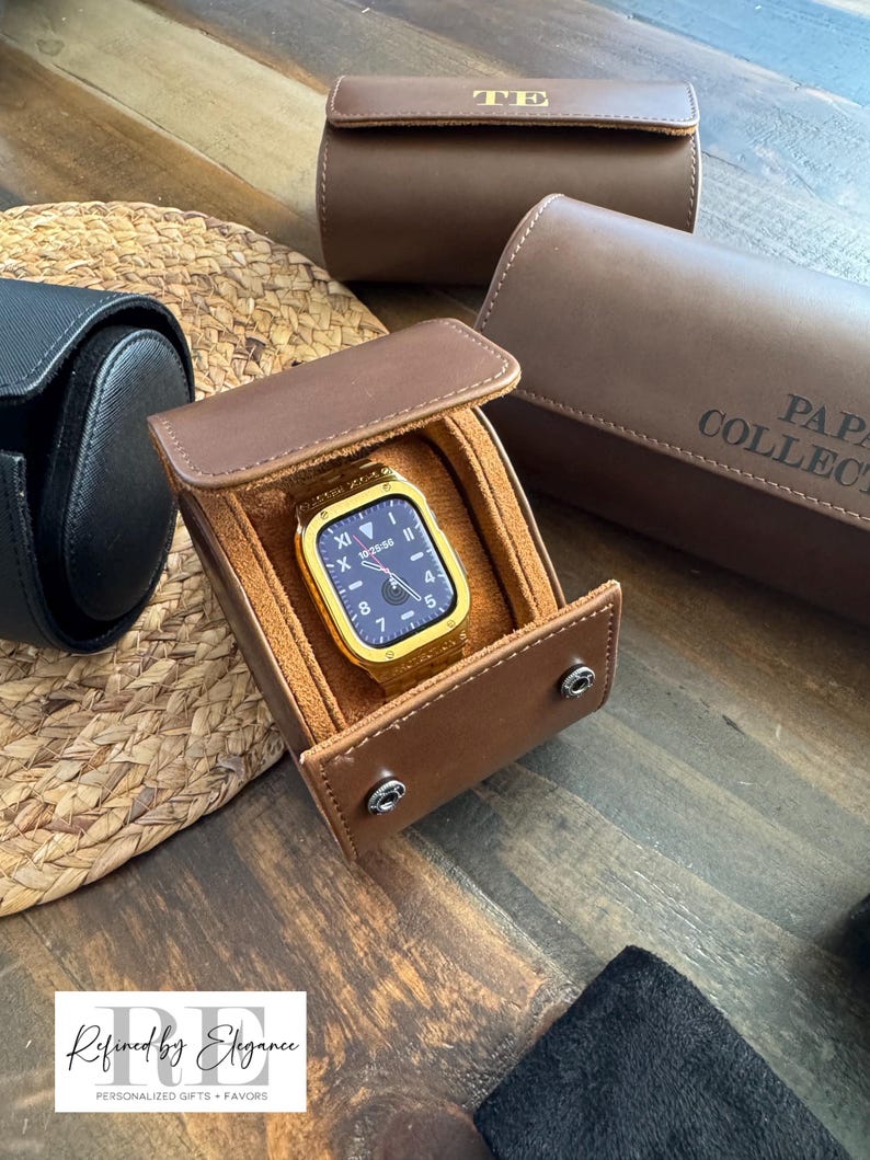 May include: A brown leather watch box with a gold watch inside. The box has a flap that closes with a snap. The watch has a black face and gold hands. The box is sitting on a wooden surface.