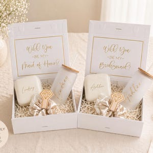 May include: Two white gift boxes with gold text that reads "Will You Be My Maid of Honor?" and "Will You Be My Bridesmaid?" Each box contains a personalized tumbler, soap, and a ring-shaped accessory. The boxes are filled with shredded paper.