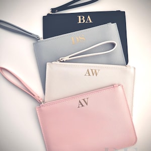 May include: Four personalized wristlet clutches in a stack. The clutches are in navy, light blue, white, and pink. Each clutch has gold initials and a matching wrist strap. The bottom right corner has the text "Refined by Elegance".