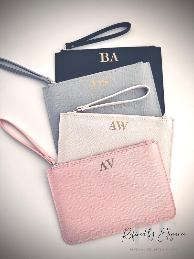 May include: Four personalized wristlet clutches in a stack. The clutches are in navy, light blue, white, and pink. Each clutch has gold initials and a matching wrist strap. The bottom right corner has the text "Refined by Elegance".