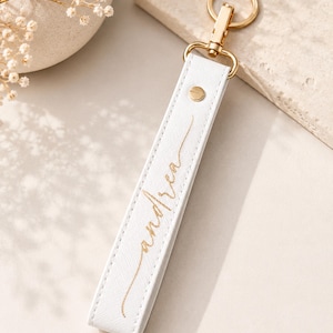 May include: A white rectangular keychain strap featuring a gold-toned metal lobster clasp and ring. The strap has a textured surface and is embossed with the name andrea in a gold cursive script, secured by a small gold-toned metal rivet.