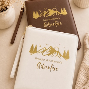 May include: Two rectangular zippered organizers with wrist straps, one dark brown and one off-white. Each features a gold foil mountain landscape graphic with birds and a sun, plus the text "Adventure" and personalized names printed in gold.