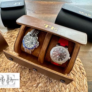 May include: A brown leather watch winder with two watch slots. The winder has a gold embossed "TE" on the front. The watch winder is sitting on a woven straw surface. Two additional black leather watch rolls are visible in the background.