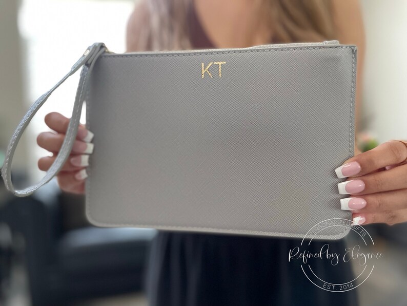 Person holding personalized grey clutch bag with gold initials.