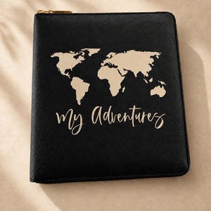 May include: A black square zippered binder featuring a gold-colored world map graphic on the front cover. Below the map, the text "My Adventures" is printed in a cursive script font. The binder has a gold-colored metal zipper pull at the top.