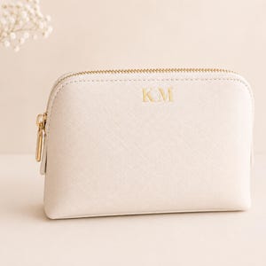 May include: A cream-colored cosmetic pouch with a textured finish and a gold-toned zipper closure. The front of the pouch features the gold-embossed letters KM centered near the top. The pouch has a structured, rounded rectangular shape.