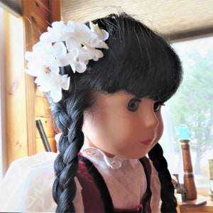 May include: A doll with long black braids and a white flower crown. She is wearing a white blouse with a red and black vest.