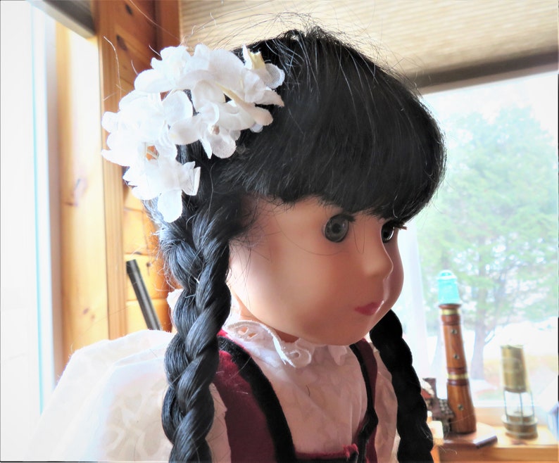May include: A doll with long black braids and a white flower crown. She is wearing a white blouse with a red and black vest.