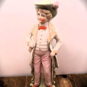 May include: A porcelain figurine of a person wearing a green hat with a pink flower, a white jacket, a pink bow tie, and pink pants. The figurine is holding a cane and standing on a green base.