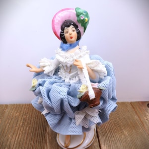 May include: A porcelain figurine of a woman in a blue dress with a pink hat. The dress has a ruffled skirt and white lace trim. The woman is holding a basket. The figurine is on a white base with gold trim.
