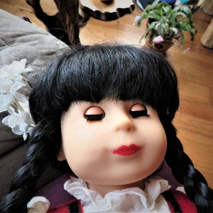 May include: A doll with black hair in braids, wearing a white blouse with a red and black vest. The doll has closed eyes and red lips. The doll is lying on a gray surface with a white flower garland in her hair.