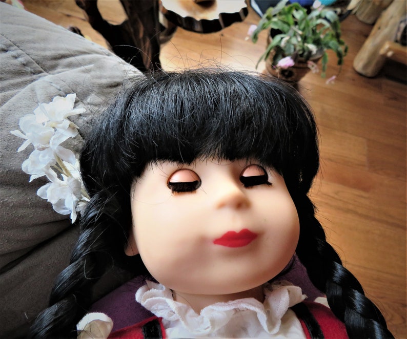 May include: A doll with black hair in braids, wearing a white blouse with a red and black vest. The doll has closed eyes and red lips. The doll is lying on a gray surface with a white flower garland in her hair.
