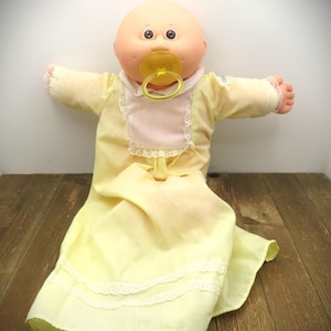 May include: A bald doll with a round peach-coloured head, dark eyes, and rosy cheeks. The doll wears a long-sleeved pale yellow nightgown featuring a white lace-trimmed bib, a white collar, and two horizontal rows of white lace trim at the hem.