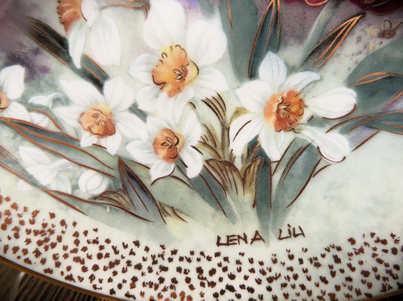 K&ouml;nnte beinhalten: Ein wei&szlig;er Porzellanteller mit einem Blumendesign mit wei&szlig;en Narzissen und gr&uuml;nen Bl&auml;ttern. Der Teller hat einen goldenen Rand und die Signatur "Lena Lih" ist unten rechts sichtbar.