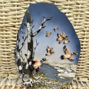 May include: A ceramic wall plaque with a winter scene featuring a birch tree and a flock of ducks flying over a frozen lake. The plaque is painted in shades of blue, brown, and white.
