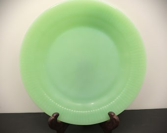 May include: A light green, round plate with a ribbed rim. The plate is made of glass and is displayed on a dark brown wooden stand. The plate has a smooth, glossy surface and a slightly raised edge.