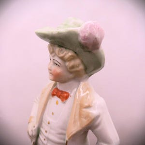 May include: A porcelain figurine of a person wearing a green hat with a pink flower, a white shirt with a red bow tie, and a tan jacket.