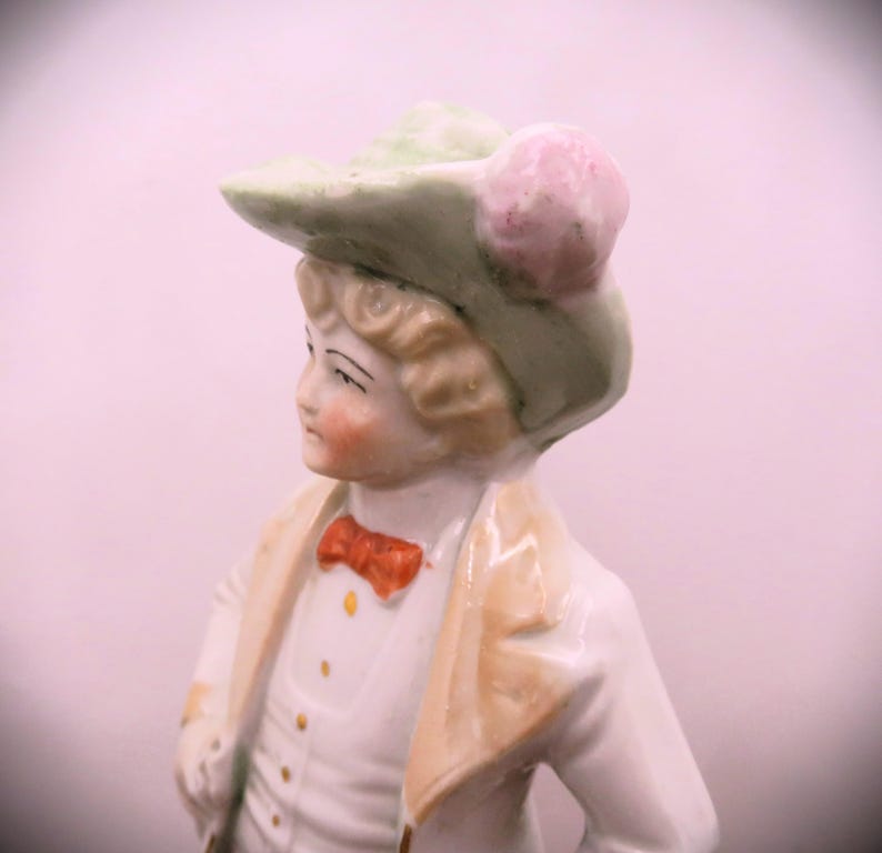 May include: A porcelain figurine of a person wearing a green hat with a pink flower, a white shirt with a red bow tie, and a tan jacket.