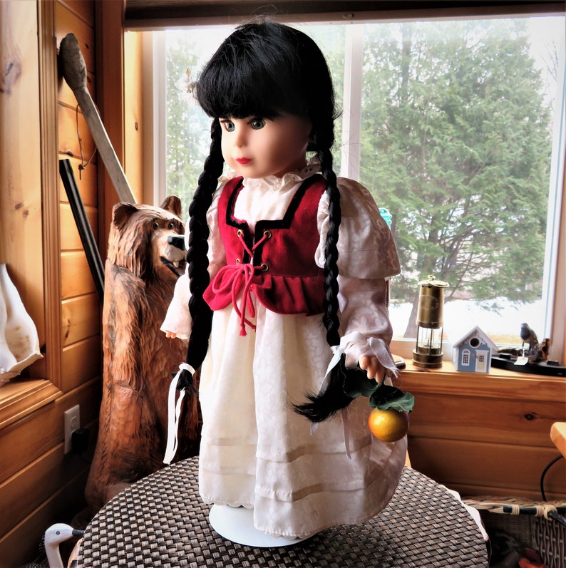 May include: A porcelain doll with black hair in braids, wearing a white dress with a red velvet vest and holding a golden apple. The doll is standing on a white pedestal.