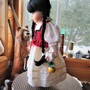May include: A porcelain doll with black hair in braids wearing a white dress with red and white trim and a red vest. The doll is holding a green leaf with a yellow fruit.