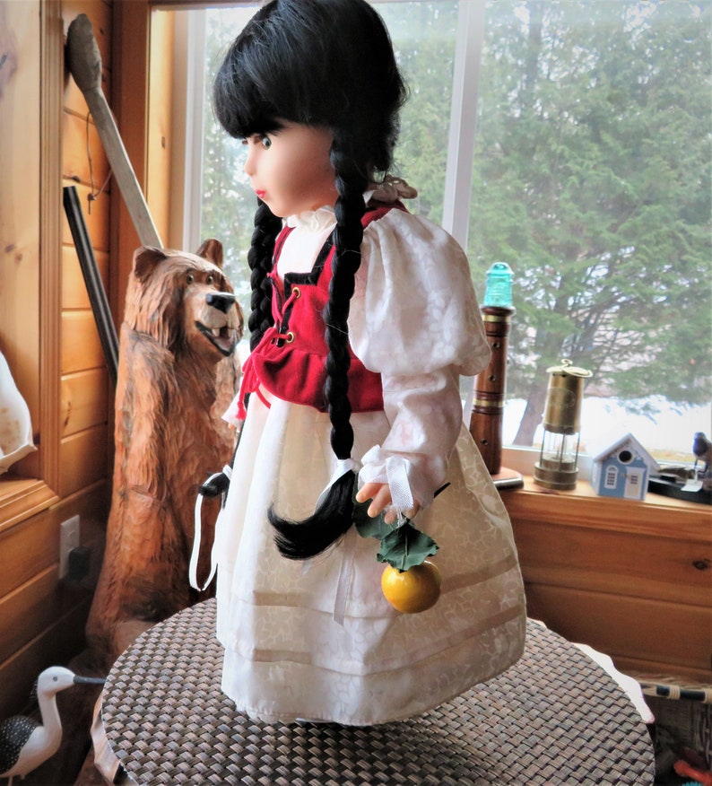 May include: A porcelain doll with black hair in braids wearing a white dress with red and white trim and a red vest. The doll is holding a green leaf with a yellow fruit.