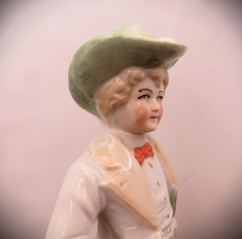 May include: A porcelain figurine of a person wearing a green hat and a white jacket with a red bow tie.