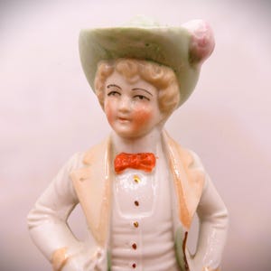 May include: A porcelain figurine of a person wearing a green hat with a pink flower, a white jacket, and a red bow tie.