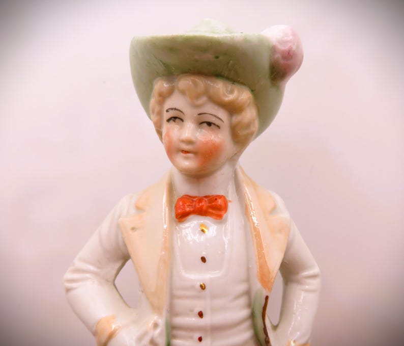 May include: A porcelain figurine of a person wearing a green hat with a pink flower, a white jacket, and a red bow tie.