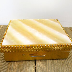 May include: A rectangular box with a gold-toned metal base and a lid featuring a smooth, cream-colored, pearlescent inlay with diagonal stripes. The lid is bordered by a decorative, textured gold-toned metal trim with a repeating beaded pattern.