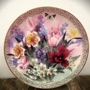 K&ouml;nnte beinhalten: Ein dekorativer Porzellanteller mit einem Blumendesign mit rosa, wei&szlig;en und roten Tulpen, gelben Narzissen und violetten Hyazinthen. Ein Schmetterling sitzt auf einer rosa Tulpe. Der Teller hat einen goldenen Rand mit einem schwarzen und goldenen gesprenkelten Muster. Der Teller ist signiert mit "Lena Liu".