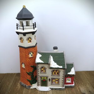 May include: A ceramic Christmas village scene featuring a tall, brick-colored lighthouse with a black top and a small house with a green roof. The house has a wreath on the door and snow accents. The scene is set on a wooden surface.