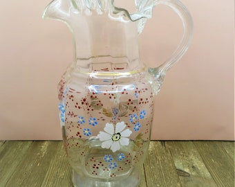 May include: A clear glass pitcher with a frilled top and a handle. The pitcher is decorated with red, blue and white painted flowers.
