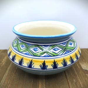 May include: A decorative ceramic bowl with a wide rim and a colorful geometric pattern. The bowl features a blue rim, a green and blue diamond pattern, and a yellow band with blue leaf-like designs. The interior is cream-colored.