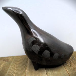May include: A dark brown wooden bird sculpture with a long neck and a rounded body. The bird has a smooth, polished finish and decorative etched details on its side. It stands on three small legs.