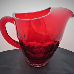 May include: A red glass pitcher with a wide mouth and a handle. The pitcher has a textured, faceted design.