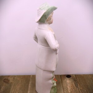 May include: A porcelain figurine of a person wearing a green hat and white clothing. The figurine is standing on a green base.