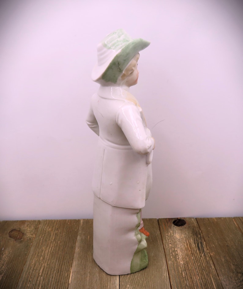 May include: A porcelain figurine of a person wearing a green hat and white clothing. The figurine is standing on a green base.