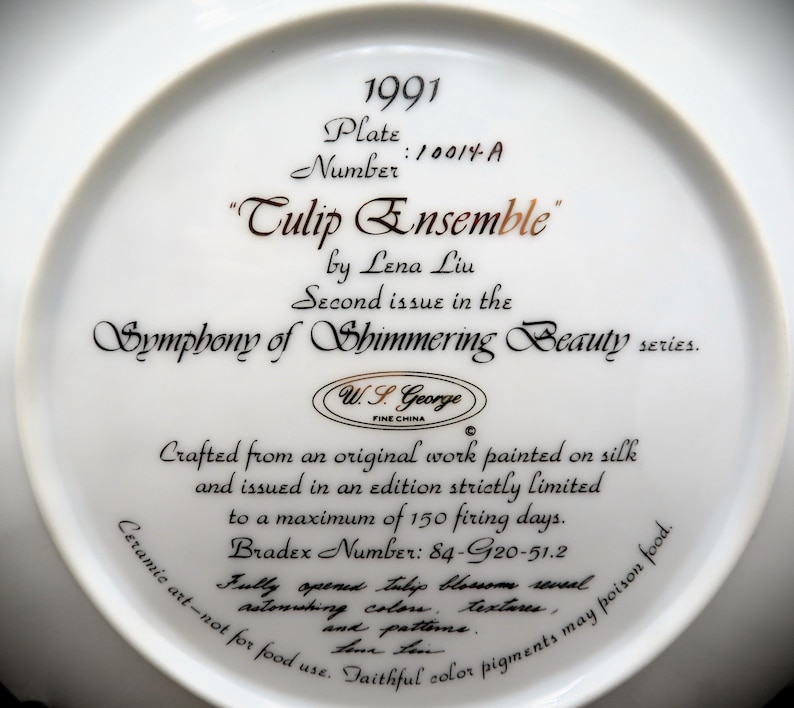 K&ouml;nnte beinhalten: Ein wei&szlig;er Keramikteller mit goldener Inschrift. Die Inschrift lautet "1991 Teller Nummer: 10014-A "Tulpenensemble" von Lena Liu Zweite Ausgabe in der Serie Symphonie der schimmernden Sch&ouml;nheit. W.S. George FEINE PORZELLAN Hergestellt aus einem Originalwerk, das auf Seide gemalt wurde, und in einer streng limitierten Auflage von maximal 150 Brenntagen herausgegeben. Bradex-Nummer: 84-G20-51.2 Die vollst&auml;ndig ge&ouml;ffnete Tulpenbl&uuml;te enth&uuml;llt erstaunliche Farben, Texturen und Muster. Keramikkunst - nicht f&uuml;r den Verzehr geeignet. Treue Farbpigmente k&ouml;nnen Lebensmittel vergiften."