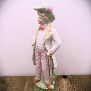 May include: A porcelain figurine of a person wearing a green hat with a pink flower, a white jacket, pink pants, and holding a cane. The figurine is standing on a green base.