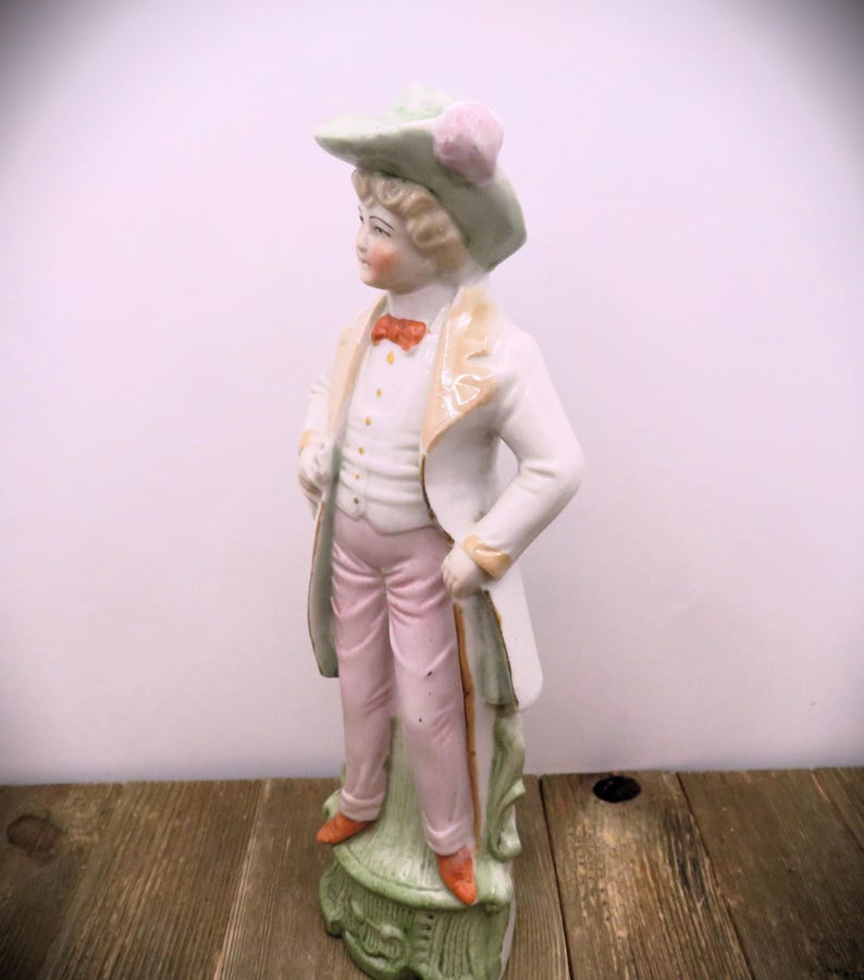 May include: A porcelain figurine of a person wearing a green hat with a pink flower, a white jacket, pink pants, and holding a cane. The figurine is standing on a green base.