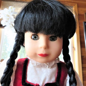 May include: A porcelain doll with black hair in braids, wearing a red and white dress with a white lace collar. The doll has blue eyes and red lips.