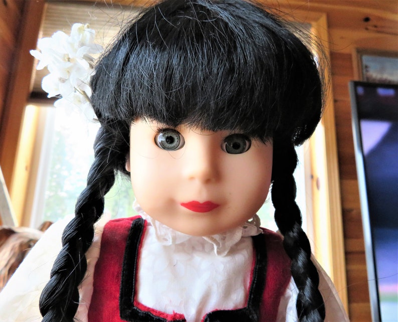 May include: A porcelain doll with black hair in braids, wearing a red and white dress with a white lace collar. The doll has blue eyes and red lips.