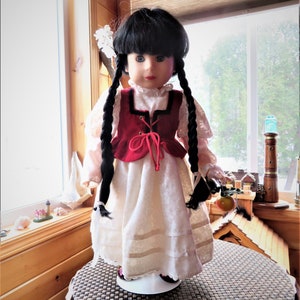 May include: A porcelain doll with black braided hair, wearing a white dress with a red vest and holding a red apple.