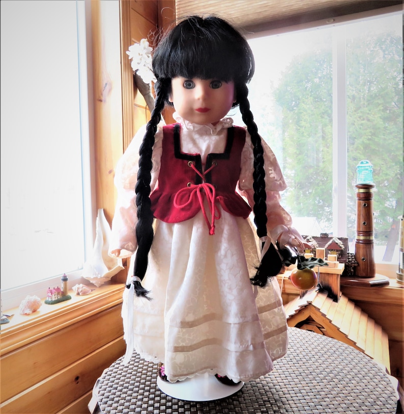 May include: A porcelain doll with black braided hair, wearing a white dress with a red vest and holding a red apple.