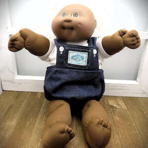 May include: A Cabbage Patch Kids doll with dark brown skin, a bald head, and brown eyes. The doll wears a white short-sleeved shirt under dark blue denim overalls featuring a rectangular white label with blue text and a floral logo on the chest.